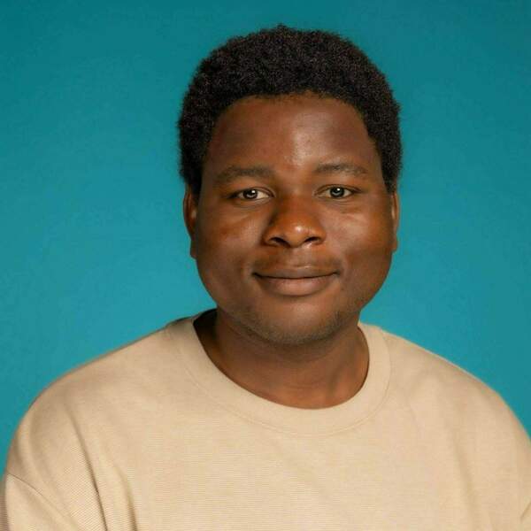 Headshot of Ikenna Nwozo, a person with short dark hair, wearing a tan shirt, against a teal background. They have a subtle smile.