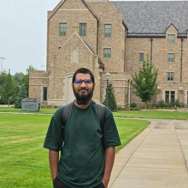 Portrait of Redwan Islam, a man with a beard and glasses wearing a dark green shirt and black pants stands in front of Morrissey Manor on a cloudy day.